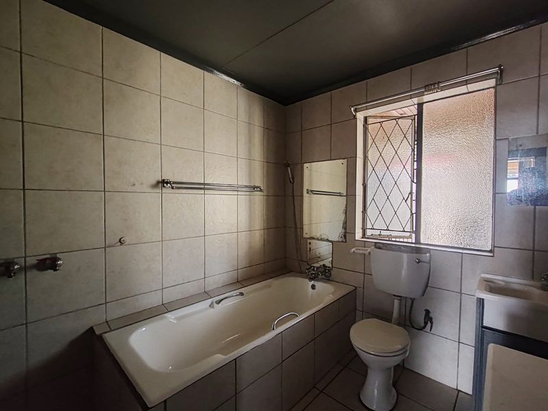 A dated bathroom featuring a built-in bathtub, toilet, and washbasin with full-height wall tiling and a security-grilled window.