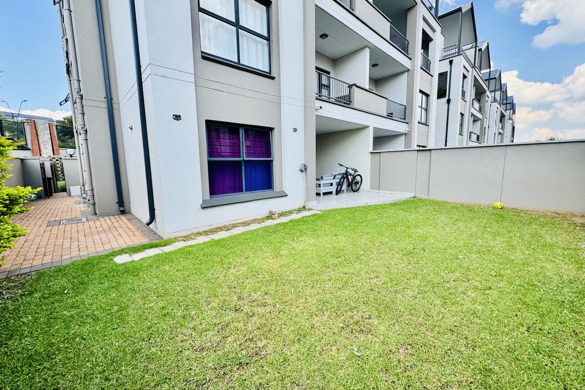 A well-maintained rear exterior view of a modern flat featuring a private lawn, paved walkway, and a covered patio area.