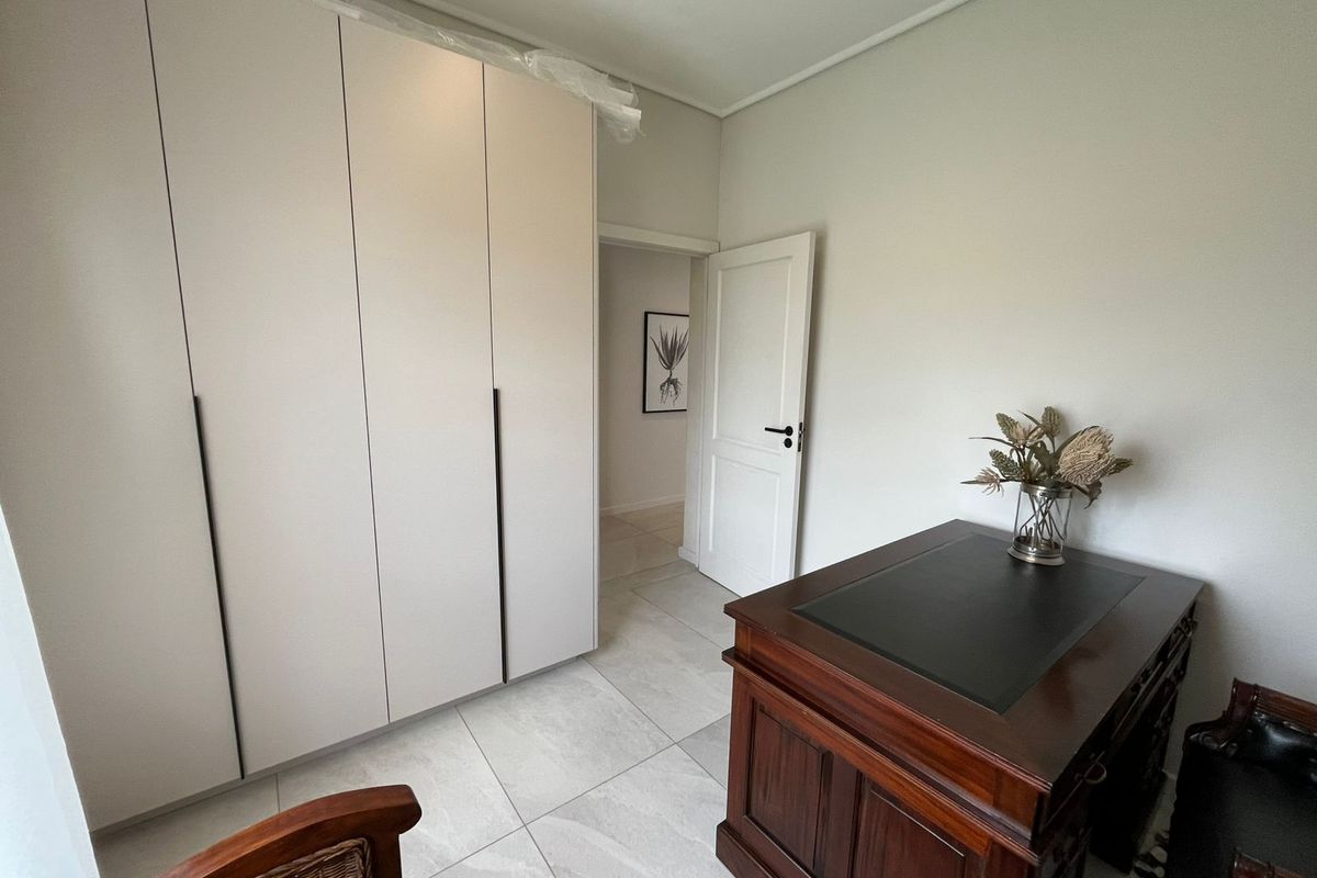 A bright bedroom featuring modern built-in floor-to-ceiling wardrobes and neutral tiled flooring, leading into a hallway.