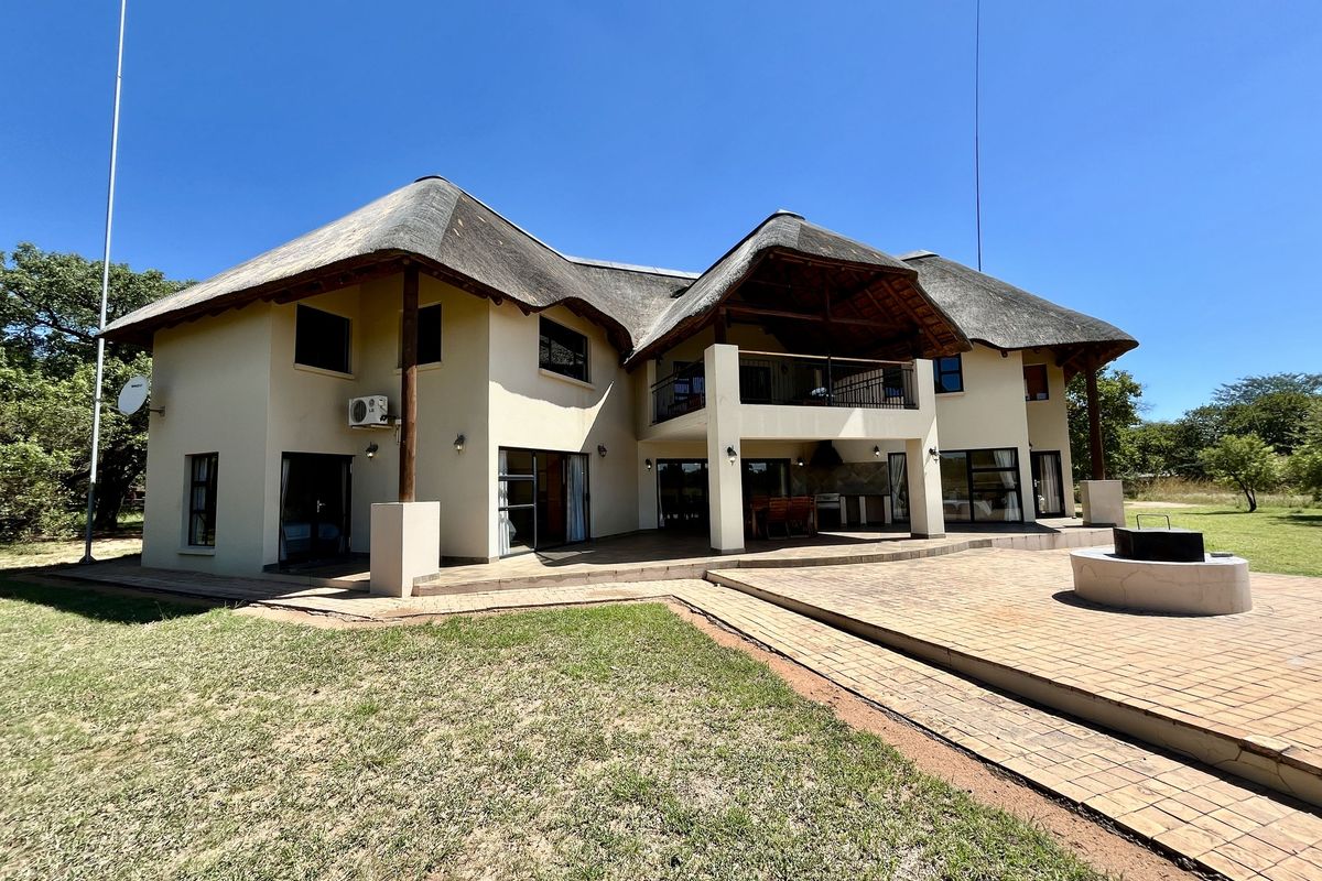 Spacious lodge exterior featuring a traditional thatched roof, expansive paved patio area, built-in fire pit, and a covered balcony overlooking the surrounding grounds.
