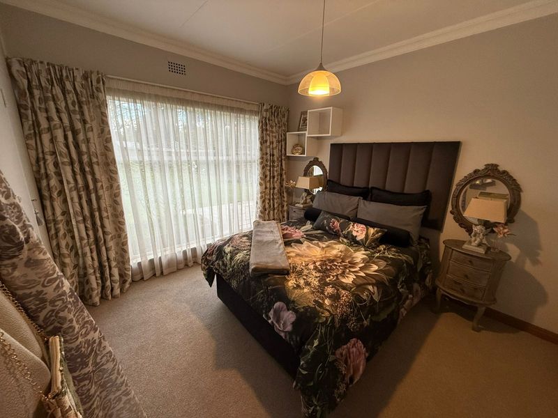 A well-maintained bedroom featuring neutral carpeting, crown molding, a large window with floor-to-ceiling curtains, and a central pendant light fixture.