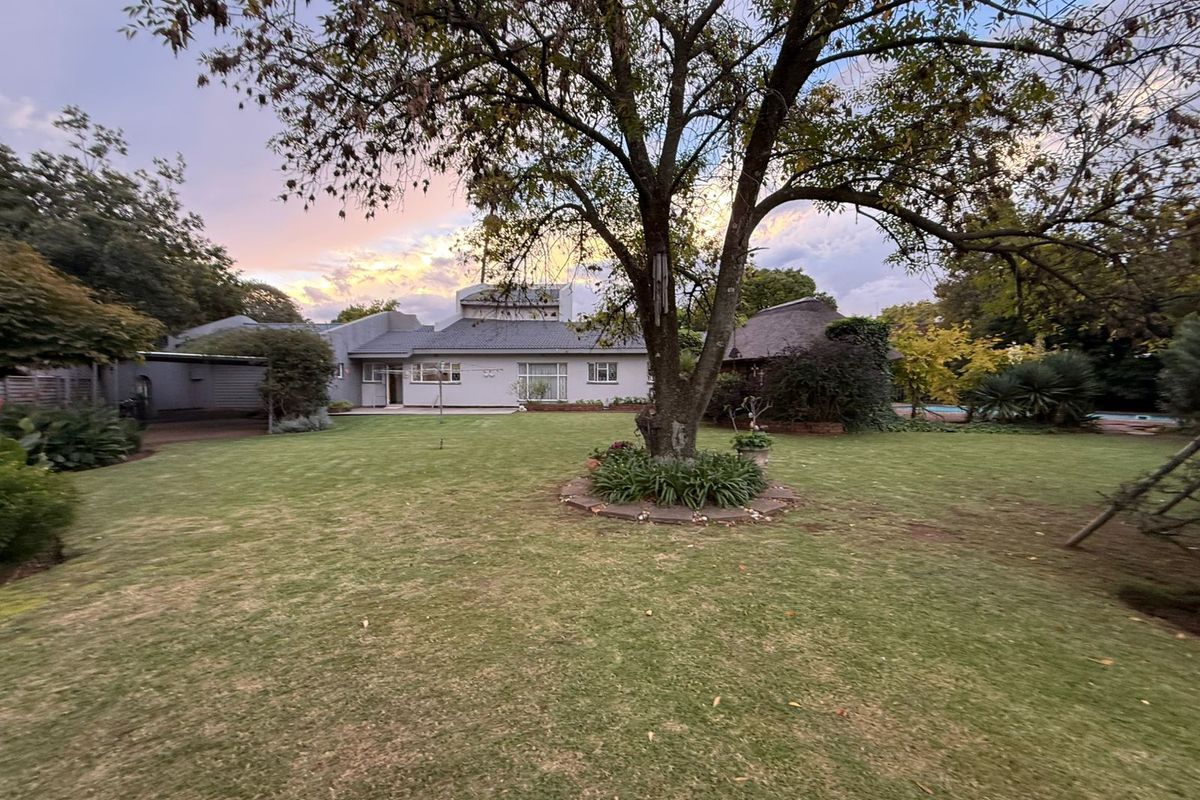 A spacious rear garden featuring a large mature tree, manicured lawn, and a view of the rear building elevation with a thatched roof structure.