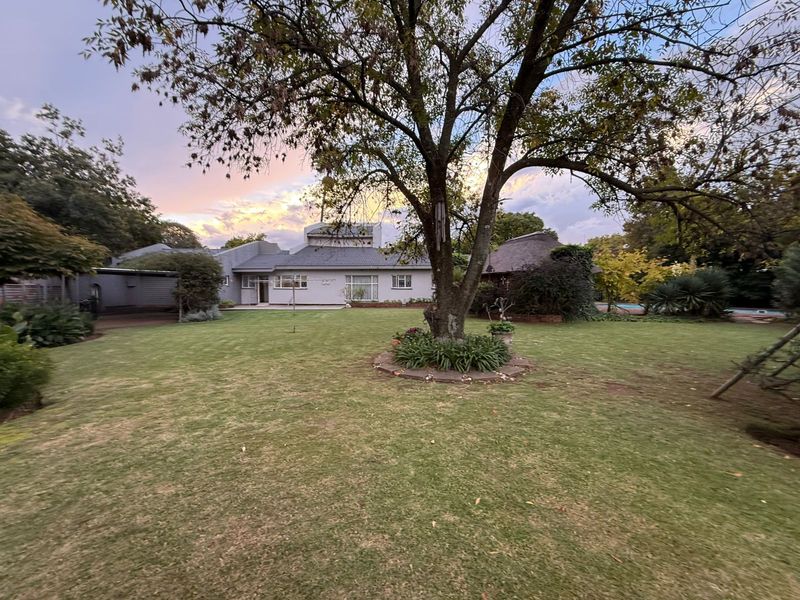 A spacious rear garden featuring a large mature tree, manicured lawn, and a view of the rear building elevation with a thatched roof structure.