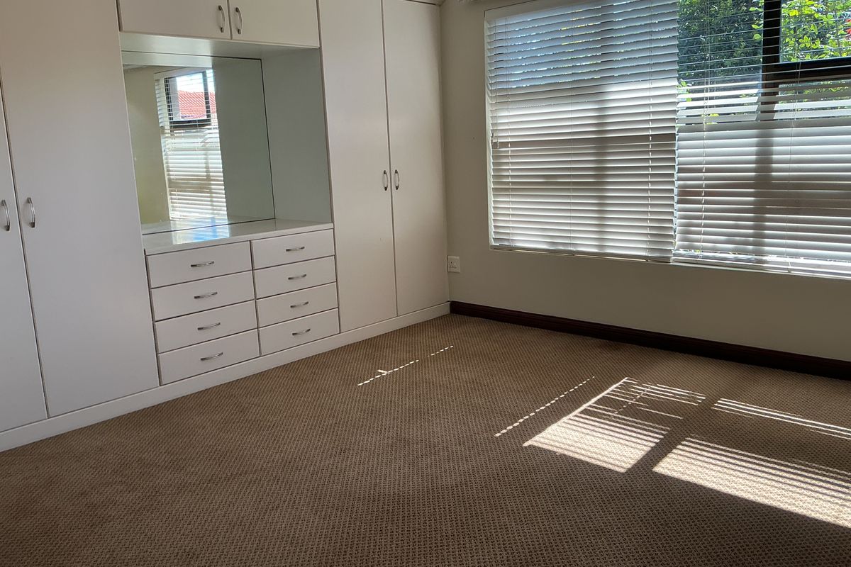A bright bedroom featuring built-in white cabinetry with integrated drawers and a mirror, complemented by neutral carpeting and window blinds.
