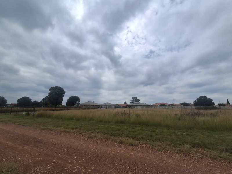 A view of a vacant residential plot featuring a dirt road access and perimeter wire fencing with surrounding neighborhood houses in the distance.