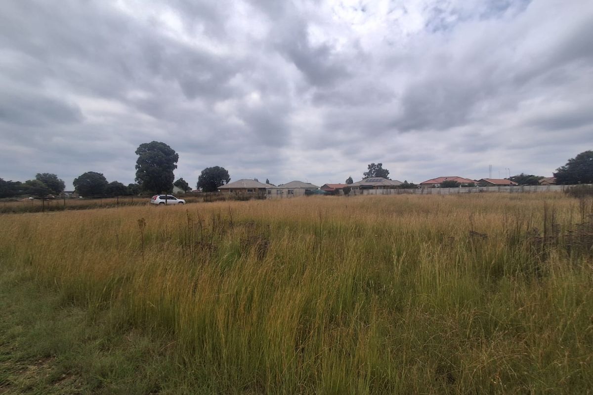 A wide-angle view of a vacant residential plot featuring tall grass and perimeter fencing, situated in a developed suburban neighborhood.