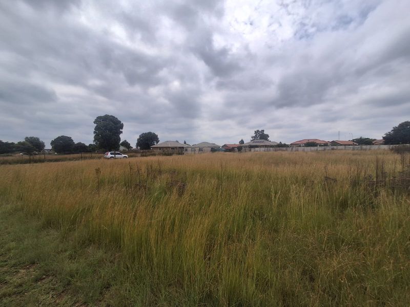 A wide-angle view of a vacant residential plot featuring tall grass and perimeter fencing, situated in a developed suburban neighborhood.