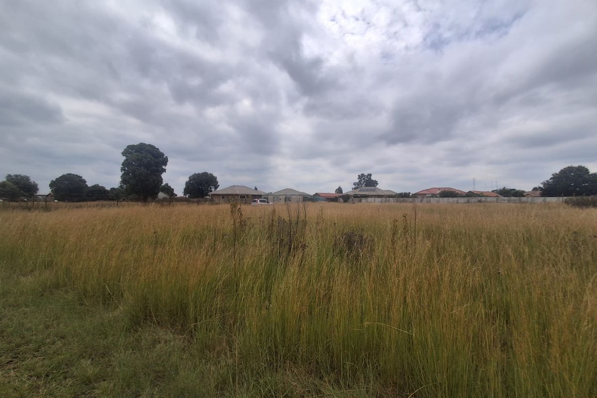 A wide-angle view of a vacant residential plot featuring tall grass and surrounding neighborhood properties in the distance.