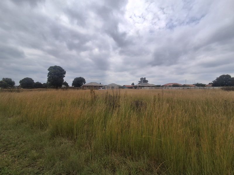 A wide-angle view of a vacant residential plot featuring tall grass and surrounding neighborhood properties in the distance.