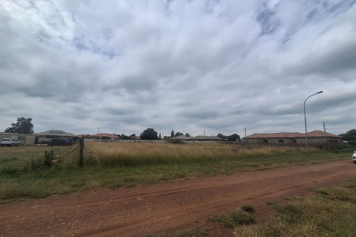 A vacant residential plot of land featuring natural grass cover, accessible via a dirt road with existing street lighting and perimeter fencing.