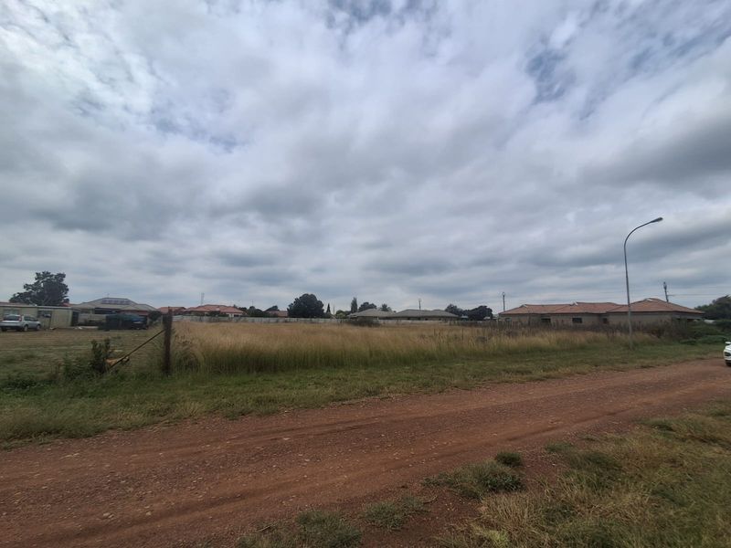 A vacant residential plot of land featuring natural grass cover, accessible via a dirt road with existing street lighting and perimeter fencing.