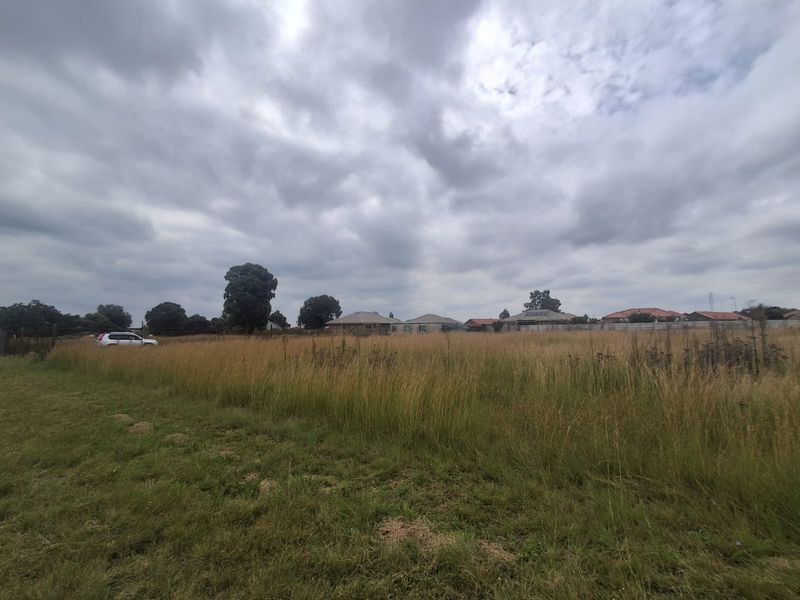 A wide-angle view of a vacant residential plot featuring open grassland with surrounding neighborhood houses visible in the background.