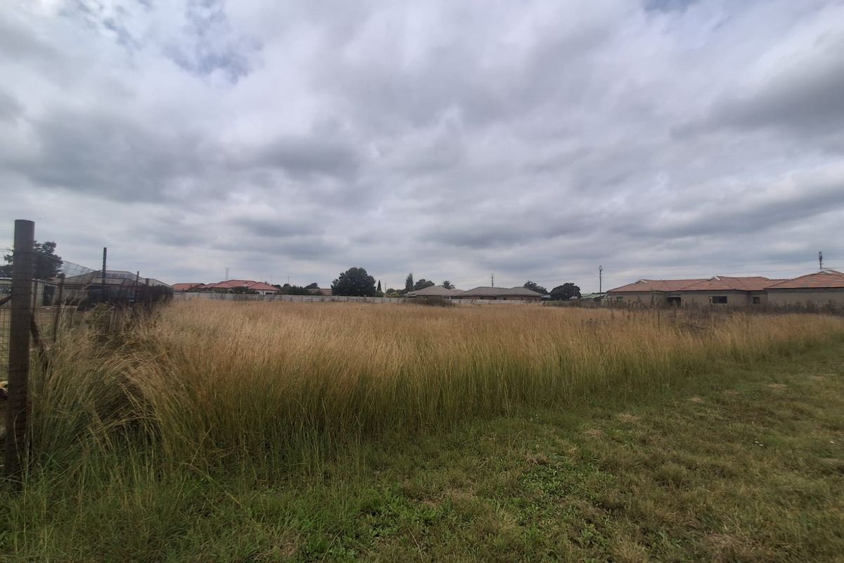 A vacant residential plot featuring overgrown grass and perimeter wire fencing, situated in a developed suburban neighborhood.
