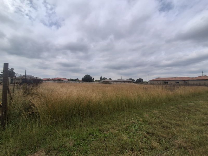 A vacant residential plot featuring overgrown grass and perimeter wire fencing, situated in a developed suburban neighborhood.