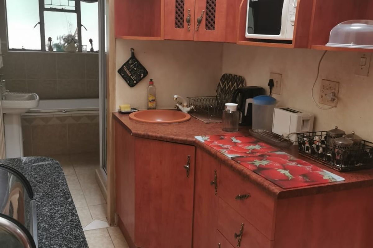 A compact kitchen area featuring dated wooden cabinetry, integrated microwave, and a small prep sink, with direct access to an adjacent bathroom.