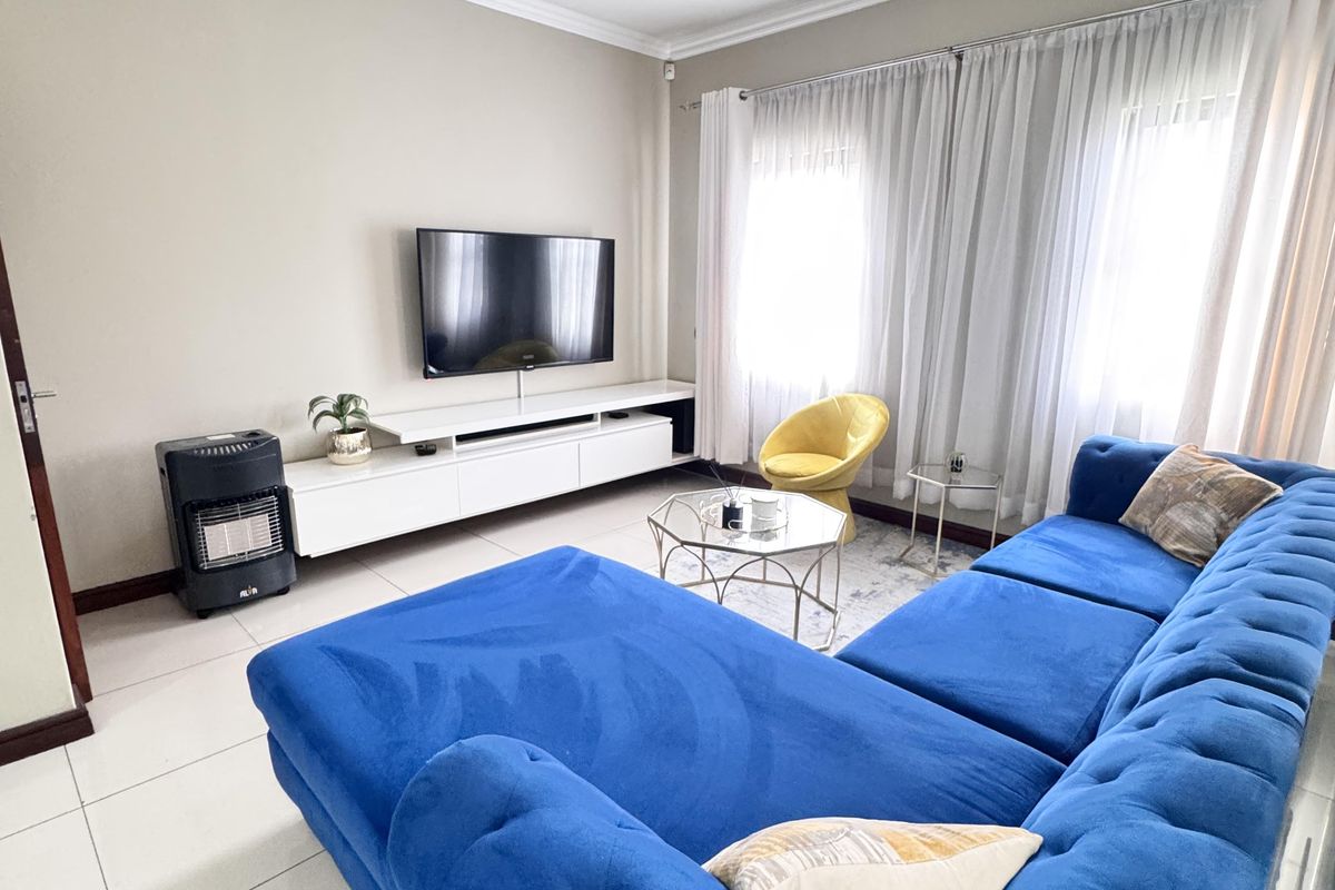 A bright and spacious living room featuring large windows with sheer curtains, tiled flooring, and a wall-mounted television unit.