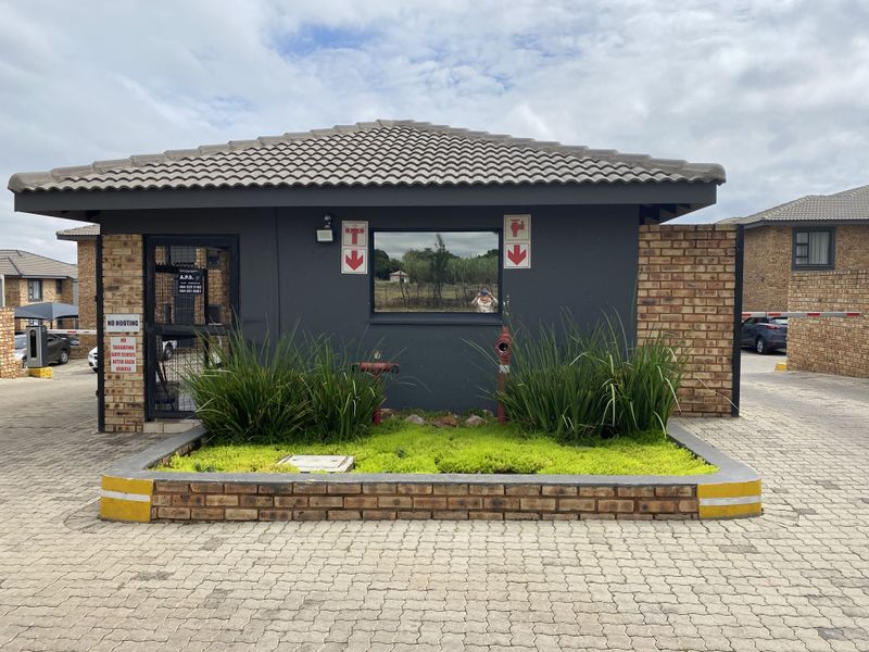 A secure gated entrance guardhouse featuring a tiled roof, brick accents, and a paved driveway approach within a townhouse complex.