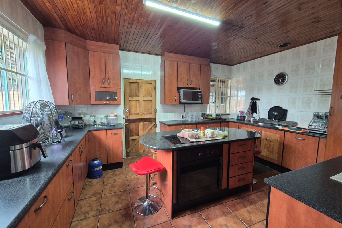 A spacious kitchen featuring wooden cabinetry, a central island with a built-in oven and hob, tiled flooring, and a distinctive wooden ceiling.