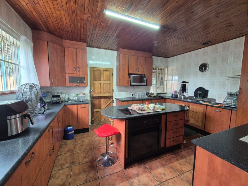 A spacious kitchen featuring wooden cabinetry, a central island with a built-in oven and hob, tiled flooring, and a distinctive wooden ceiling.