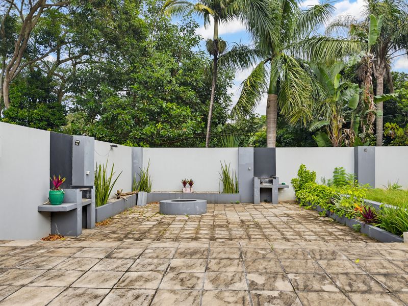 Outdoor entertainment area featuring a paved patio, built-in braai stations, and a central fire pit, set against a landscaped garden backdrop.