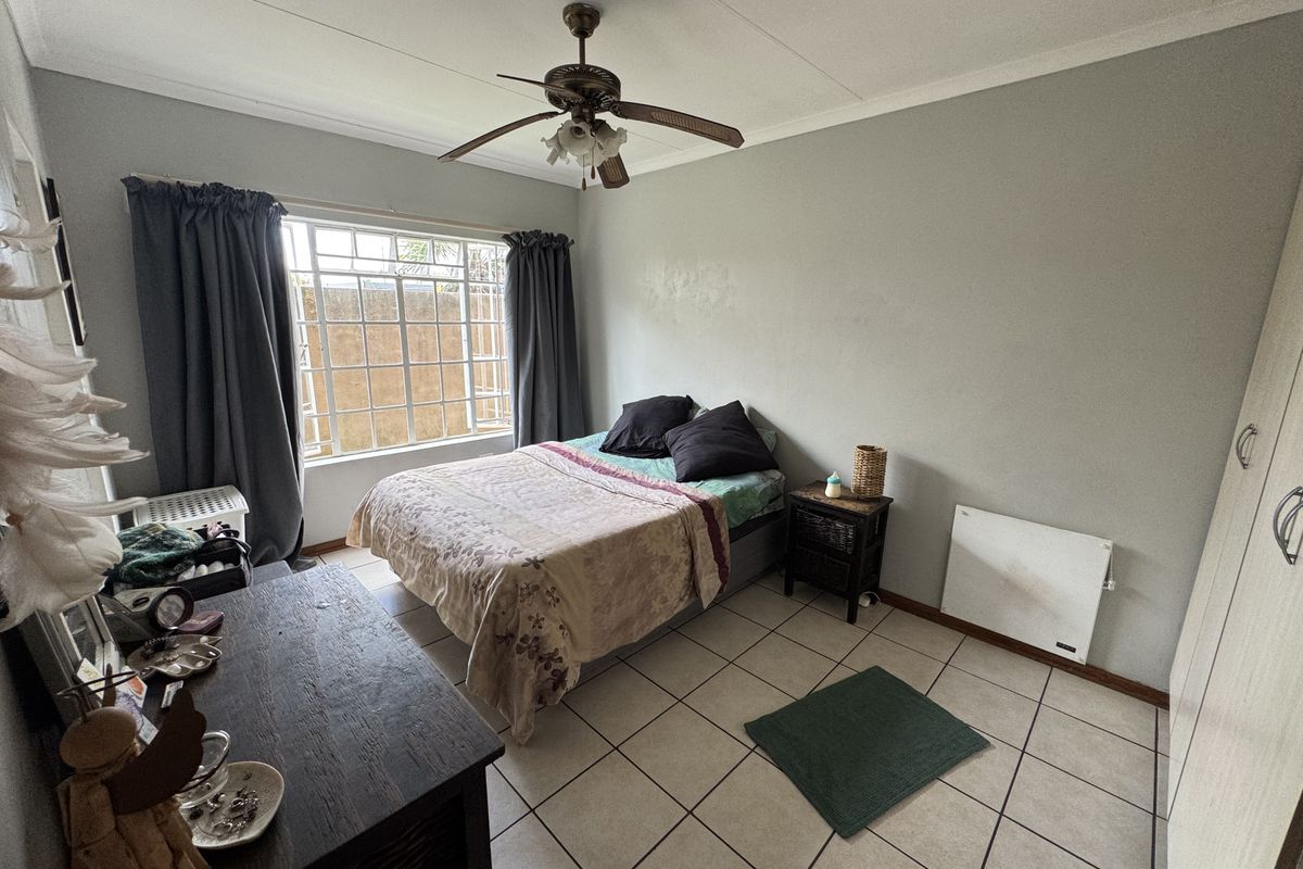 A functional bedroom featuring tiled flooring, a ceiling fan, built-in cupboards, and a large window with security bars.
