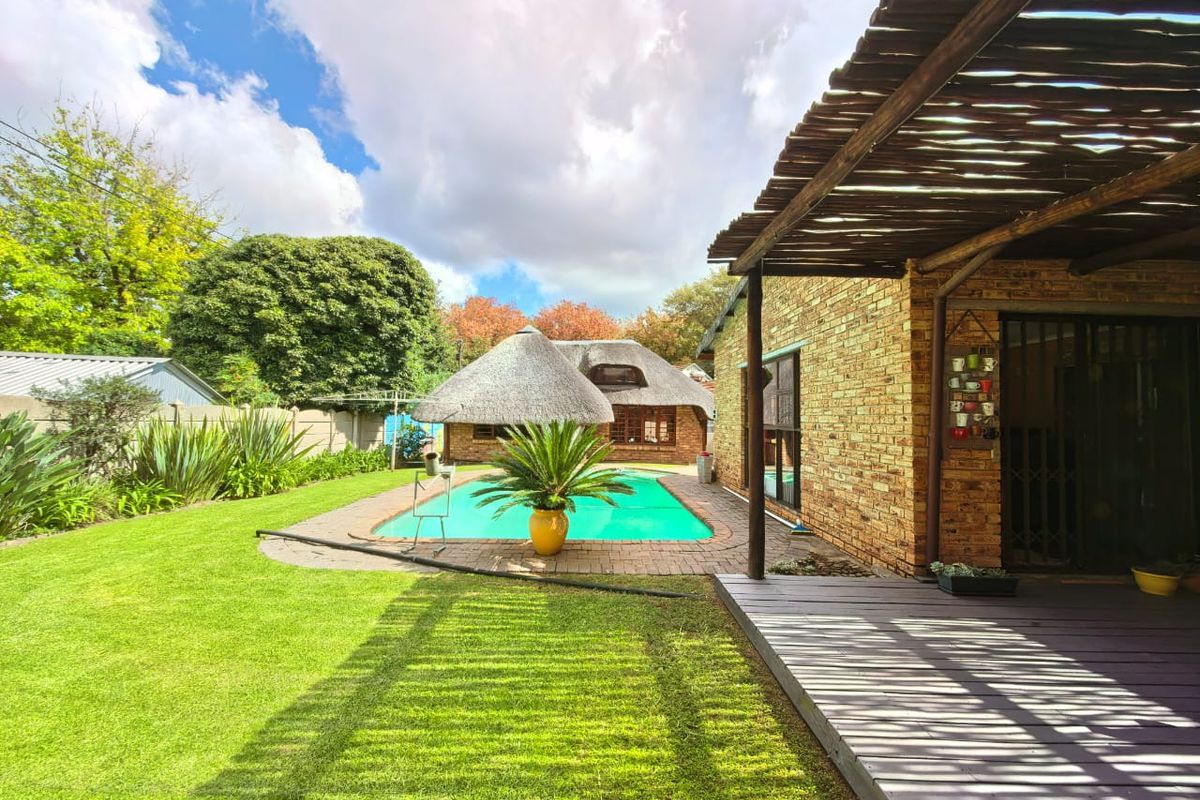 A well-maintained backyard featuring a swimming pool, a thatched lapa, a wooden deck, and a brick-clad exterior wall.