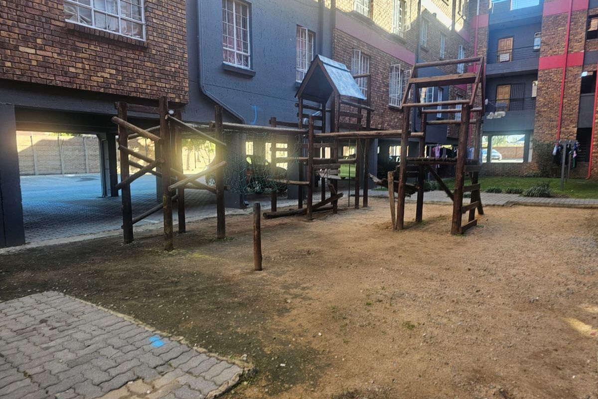 Communal outdoor area featuring a wooden playground structure situated between townhouse blocks with paved pathways and sandy ground.