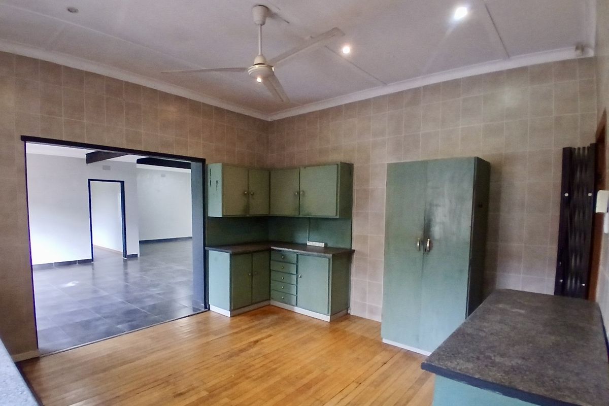 A dated kitchen area featuring built-in cabinetry, tiled walls, and timber flooring, with an open-plan flow into the adjacent living space.