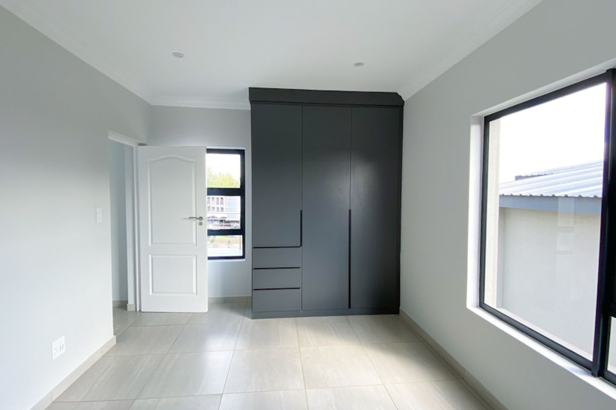 A bright, modern bedroom featuring neutral tiled flooring, crisp white walls, and a sleek, dark grey built-in wardrobe.