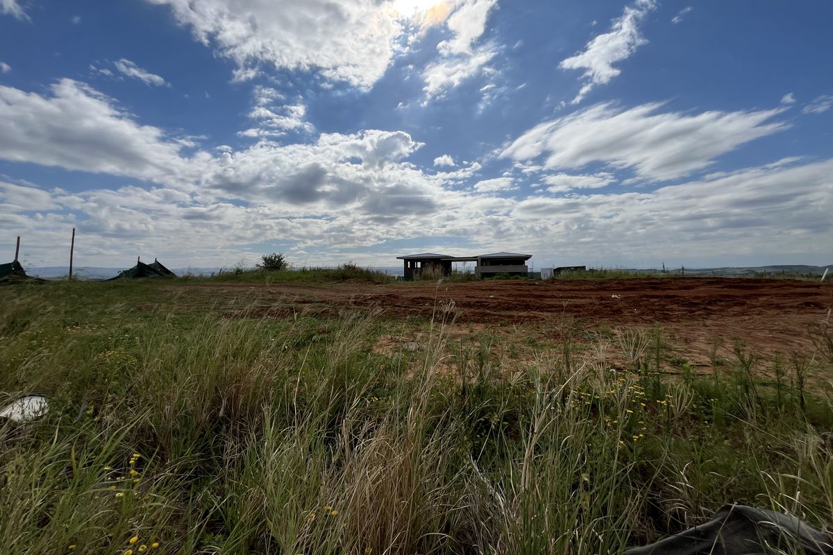 A wide-angle view of a vacant residential plot featuring cleared earth and distant structures under a bright, cloudy sky.