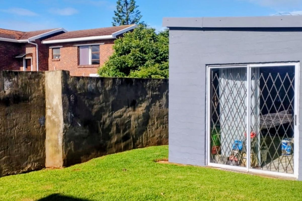 A rear garden area featuring a grey painted outbuilding with sliding glass doors and security gates, adjacent to a high boundary wall.