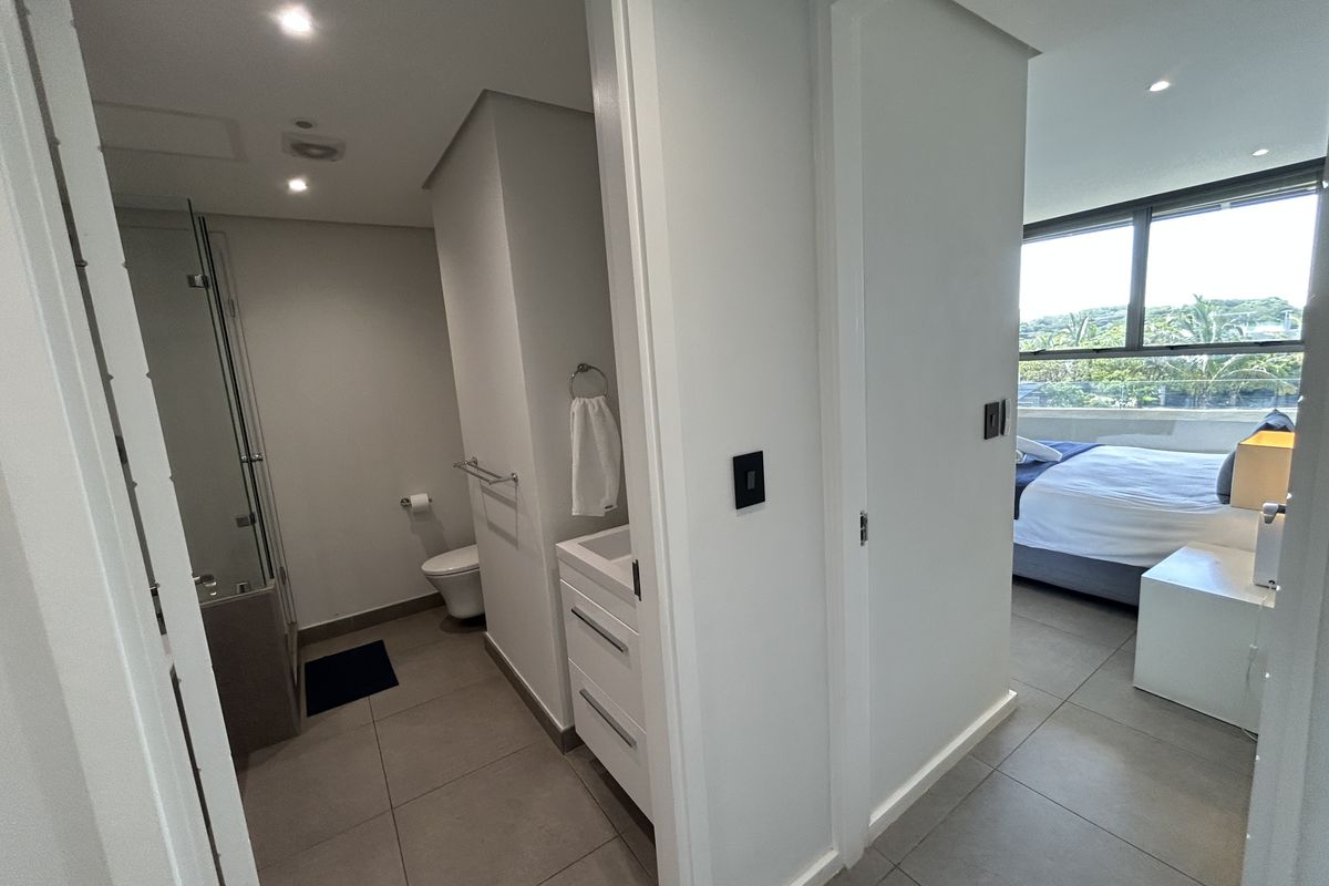 A modern bathroom featuring a vanity cabinet, wall-mounted toilet, and glass shower enclosure, adjacent to a bedroom with large windows.