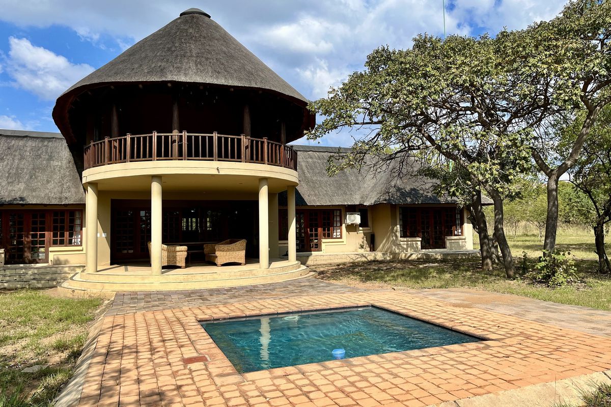 A rear view of a thatched-roof house featuring a covered patio, a swimming pool, and a spacious garden area.