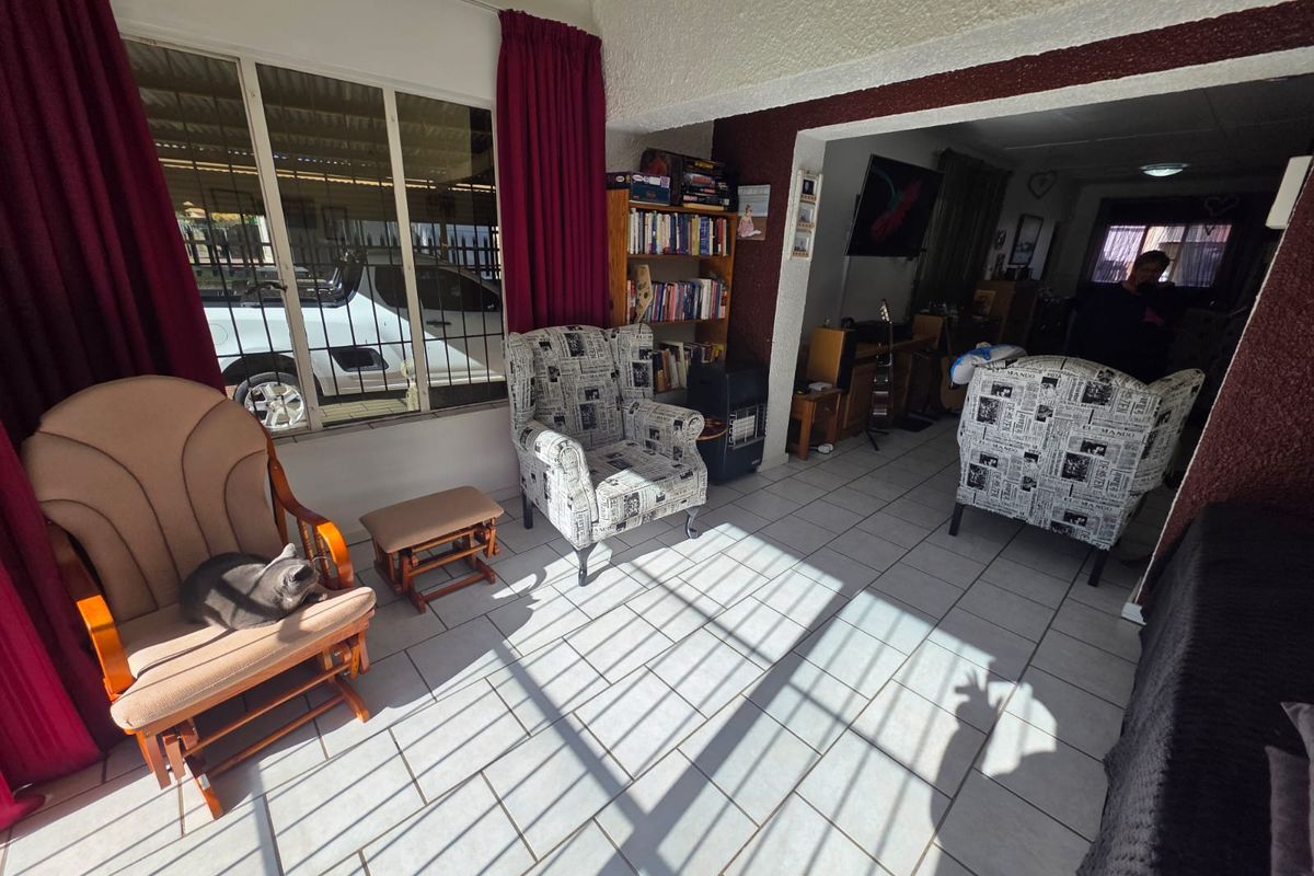 A sunlit living area featuring tiled flooring, large windows with security bars, and an open-plan layout connecting to the adjacent room.