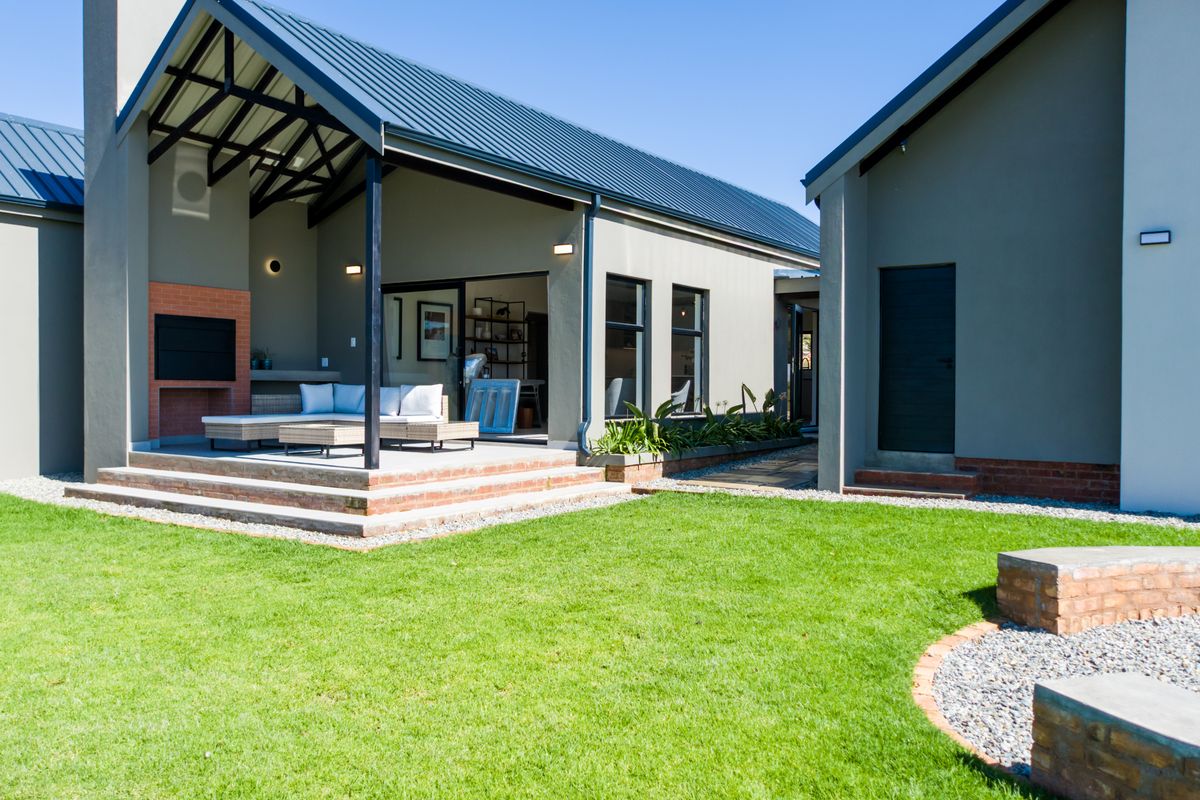 A modern rear exterior featuring a covered patio with a built-in braai, brick steps, and a landscaped garden with a brick fire pit.