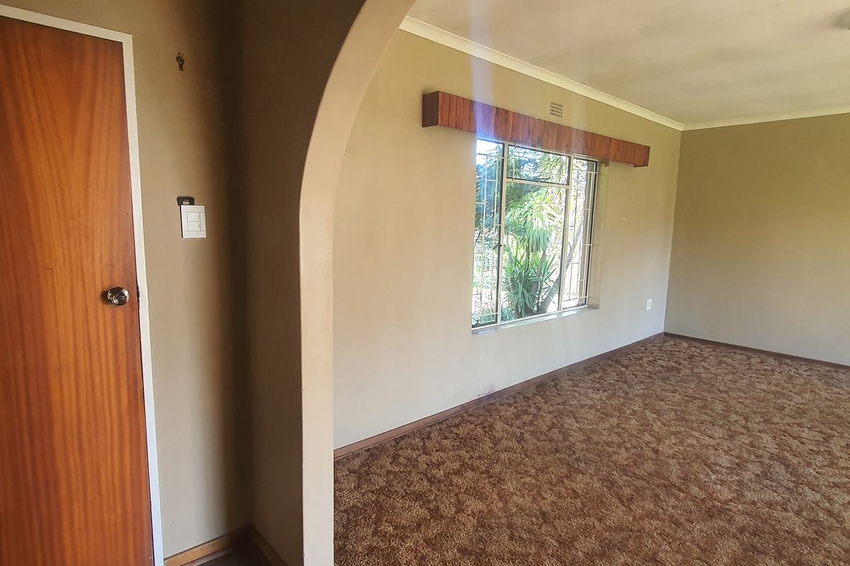 An empty living room featuring an arched entryway, large window with a wooden pelmet, and dated wall-to-wall carpeting.
