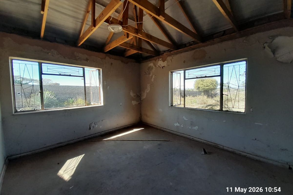 An unfinished interior room featuring exposed timber roof trusses, concrete flooring, and large windows with security bars, requiring significant renovation and finishing work.