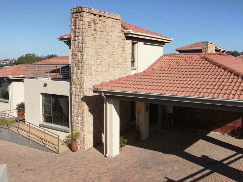 Elevated view of the property facade featuring a stone-clad chimney, terracotta roof tiles, paved driveway, covered carport, and an accessible wheelchair ramp.
