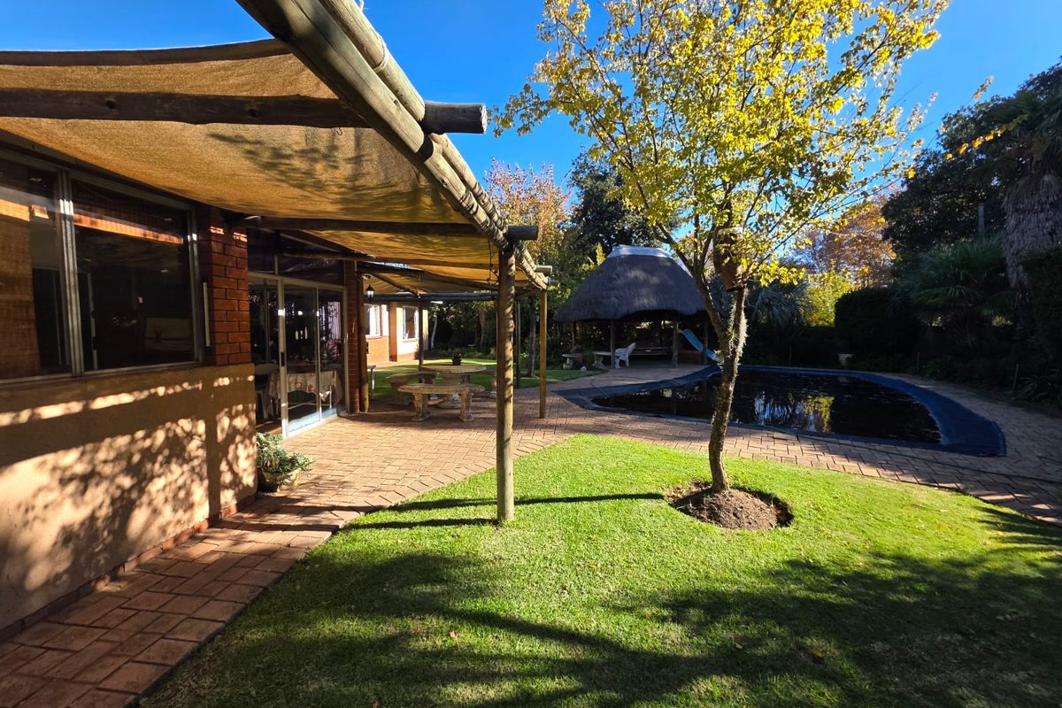 Rear garden area featuring a paved patio with shade sail, swimming pool, and a traditional thatched lapa structure.
