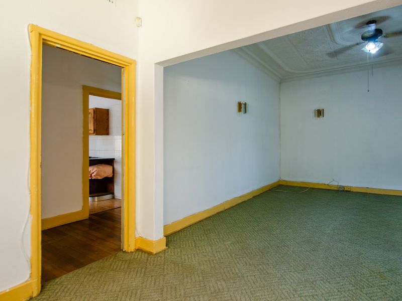 An empty living room featuring original ceiling molding, a ceiling fan, and green carpeting, with an adjacent view into a kitchen area.