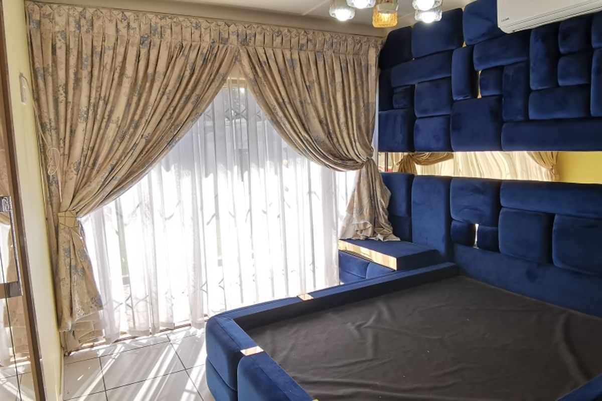 A well-maintained bedroom featuring a custom blue upholstered headboard wall, tiled flooring, and sliding glass doors with curtains leading to an outdoor area.