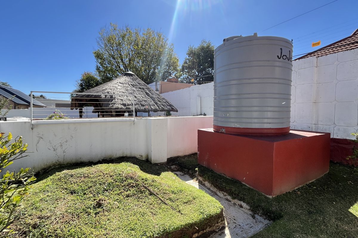 Rear garden area featuring a raised water storage tank on a concrete plinth, perimeter boundary walls, and a thatched lapa structure in the background.