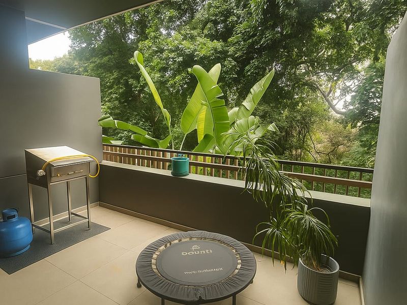 A covered balcony featuring tiled flooring, a built-in gas braai station, and a wooden balustrade overlooking lush greenery.