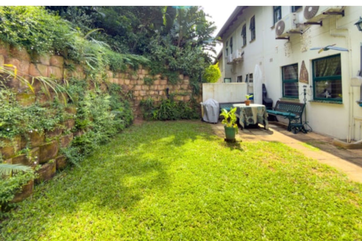 A private rear garden featuring a lawn, paved patio area, and a stone retaining wall with established greenery.