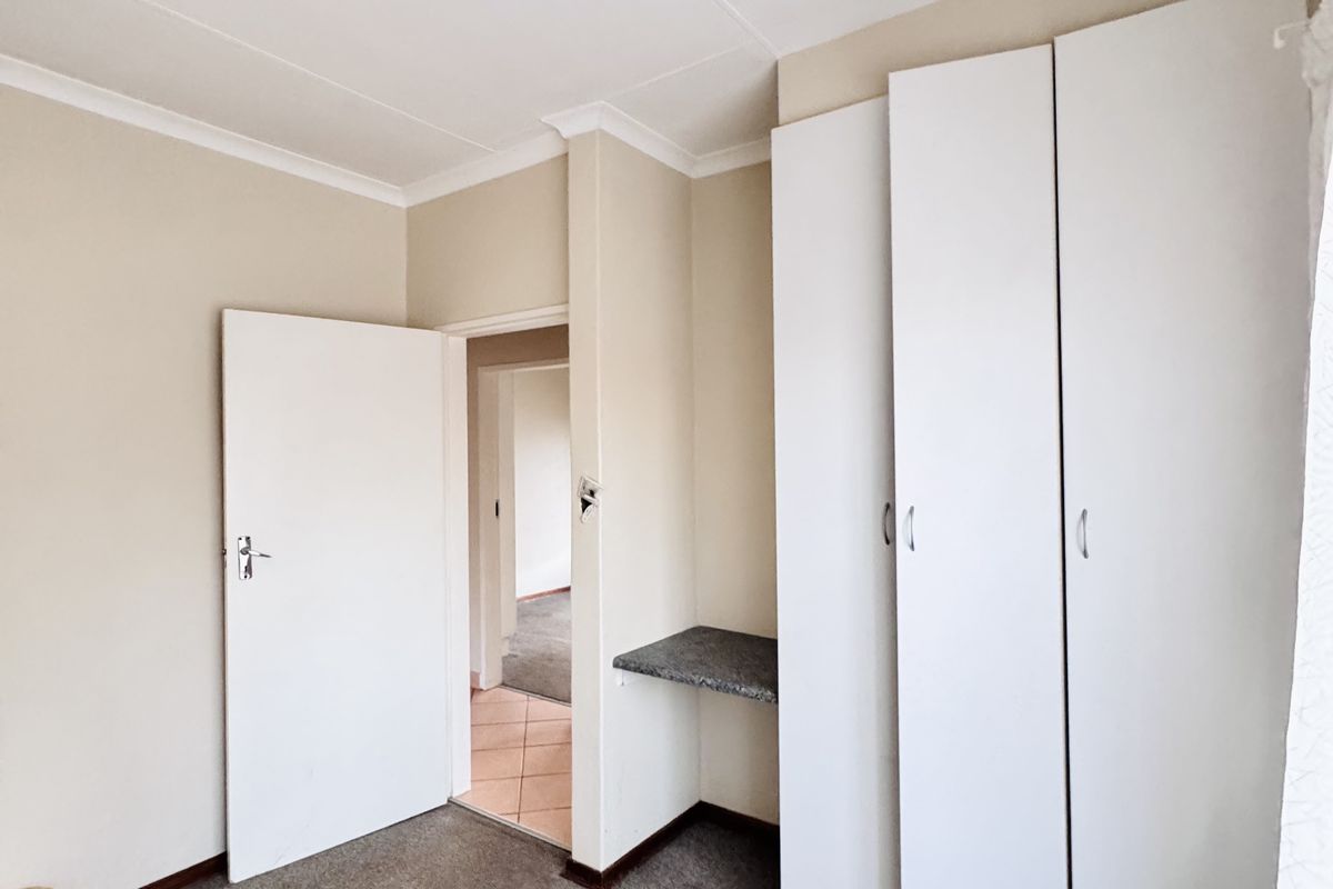 A bedroom featuring built-in cupboards, a small integrated desk area, and carpeted flooring, leading into an adjacent hallway.