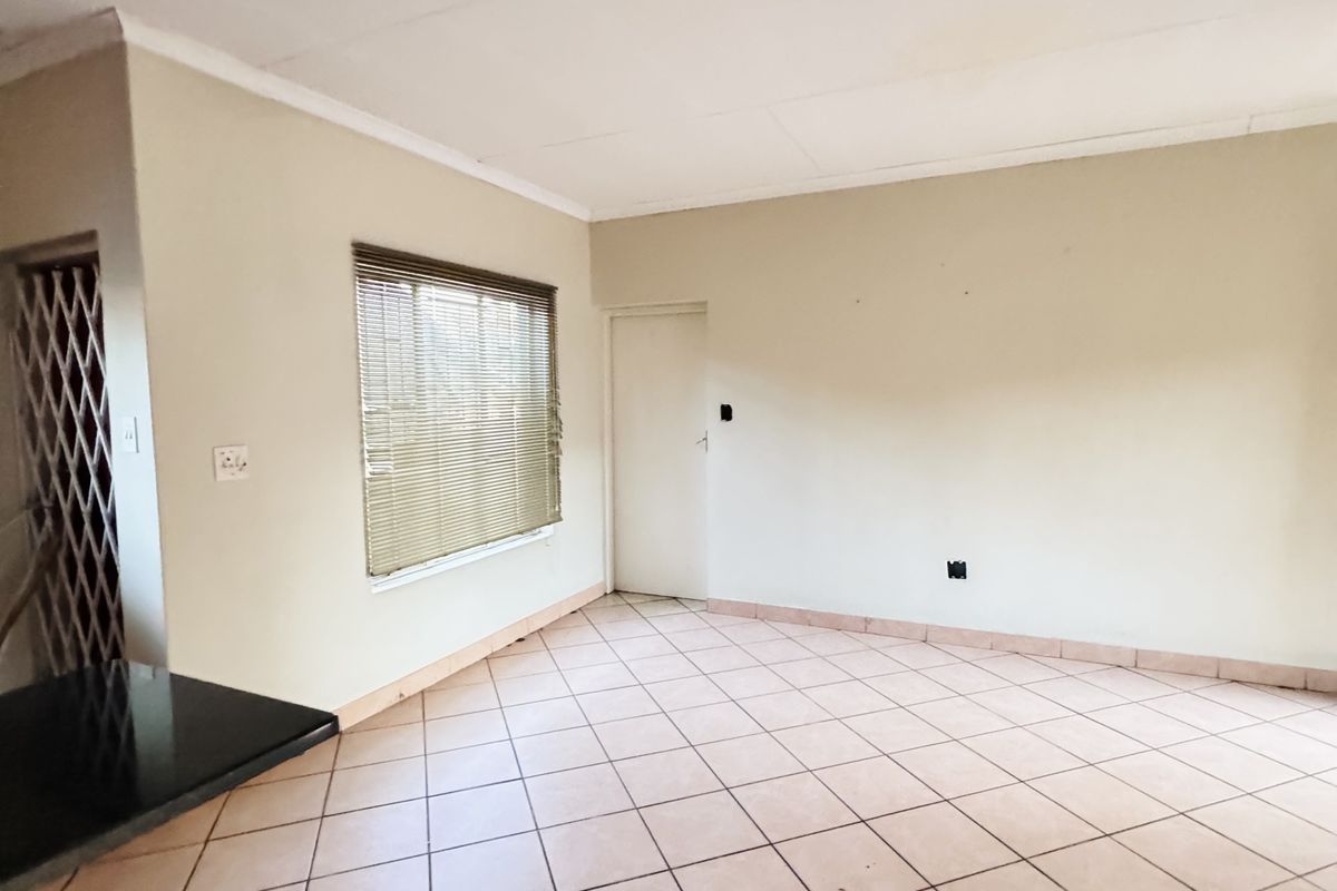 A spacious, empty living area featuring neutral tiled flooring, a window with fitted blinds, and a security gate at the entrance.