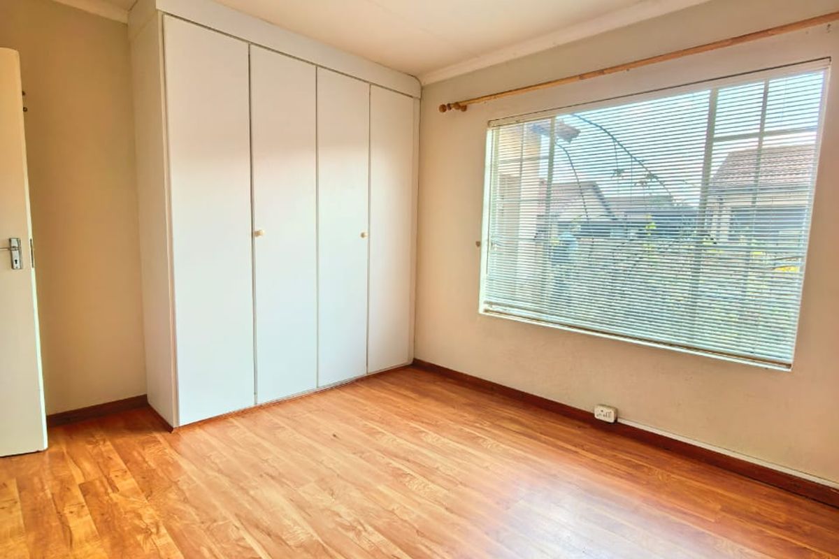 A bright, empty bedroom featuring laminate flooring, built-in cupboards, and large windows with fitted venetian blinds.