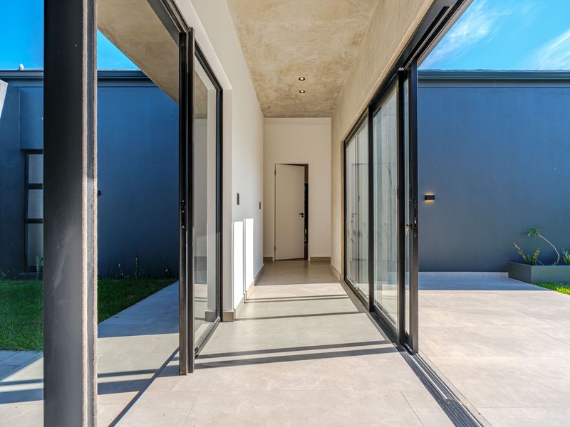 A modern covered walkway featuring large sliding glass doors, tiled flooring, and a raw concrete ceiling, connecting outdoor living spaces.