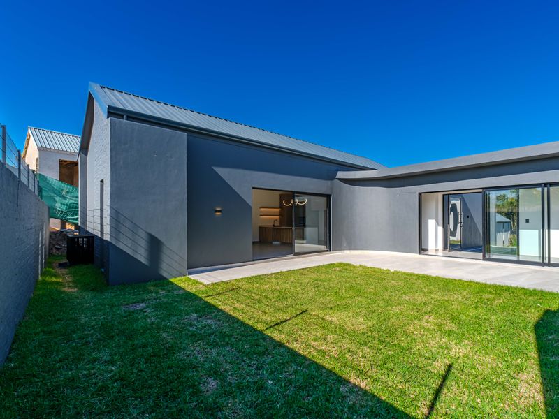 Modern rear exterior view featuring a dark grey facade, metal roof, paved patio area, and sliding glass doors leading into the home.