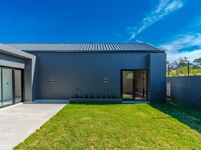 Modern rear exterior view featuring a dark grey facade, metal roof, paved patio area, and a well-maintained lawn with a built-in planter.